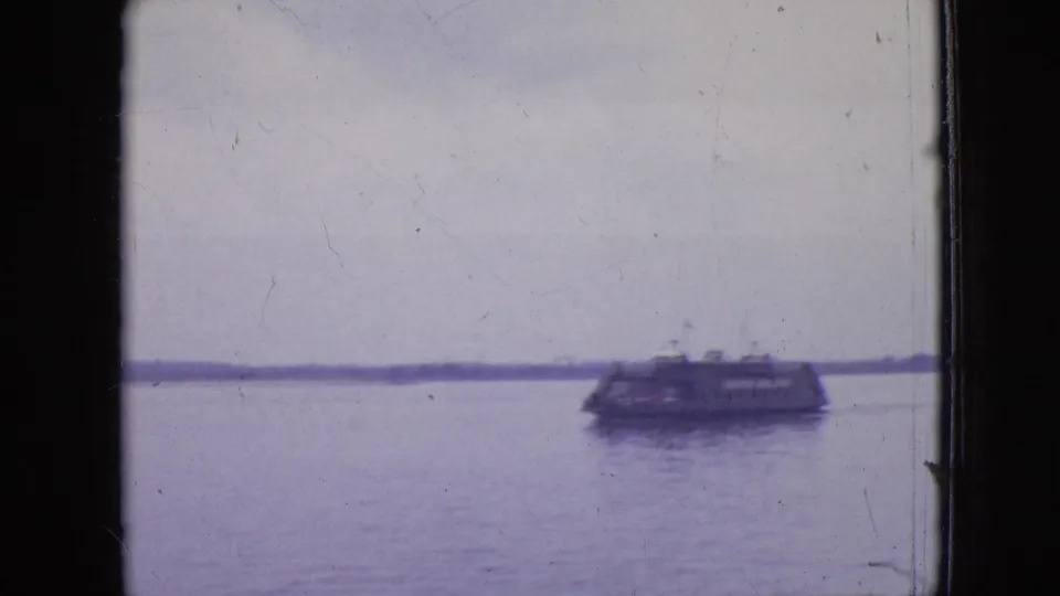 1964 Philadelphia home-movie frame with a distant ferry or barge crossing calm water under an overcast sky, with faint lettering on the vessel.