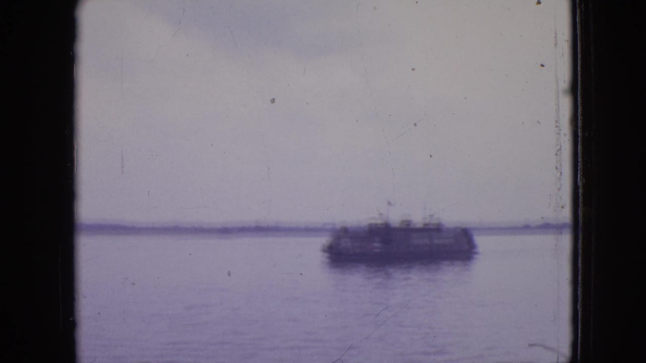 A distant sightseeing or ferry-style boat crosses calm water in 1964 Philadelphia home-movie footage, seen through a dark vertical frame edge with a low shoreline beyond.
