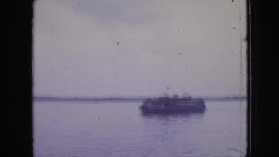 A distant sightseeing or ferry-style boat crosses calm water in 1964 Philadelphia home-movie footage, seen through a dark vertical frame edge with a low shoreline beyond.