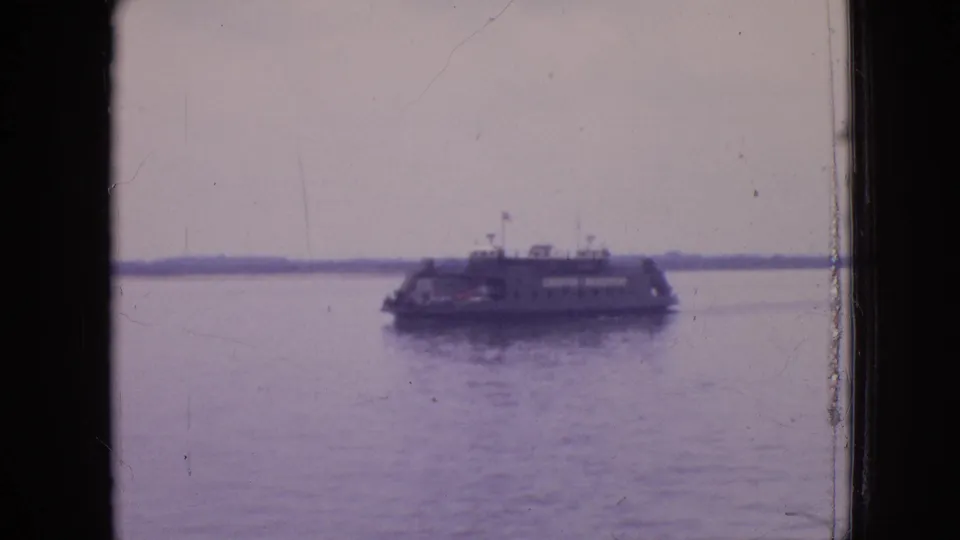 The boat sits near the right side of the frame in the 1964 Philadelphia home movie, with calm water, faint reflection, and a low distant shoreline under an overcast sky.