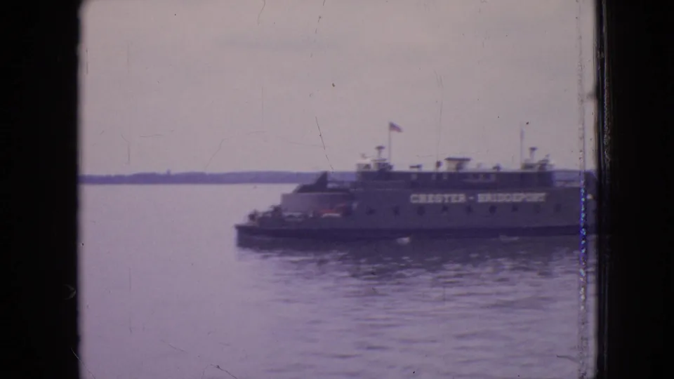 1964 8mm home-movie frame showing the Chester-Bridgeport ferry on calm water, with a low shoreline and dark framing from a window or film gate.