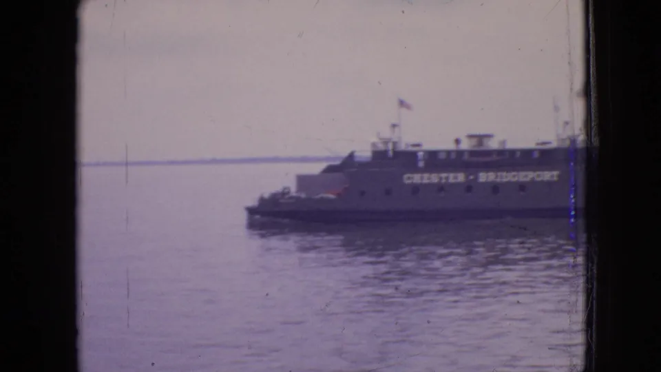 The ferry remains in view near the right side of the frame, with Chester-Bridgeport lettering visible through soft, scratched 8mm footage and calm water below.