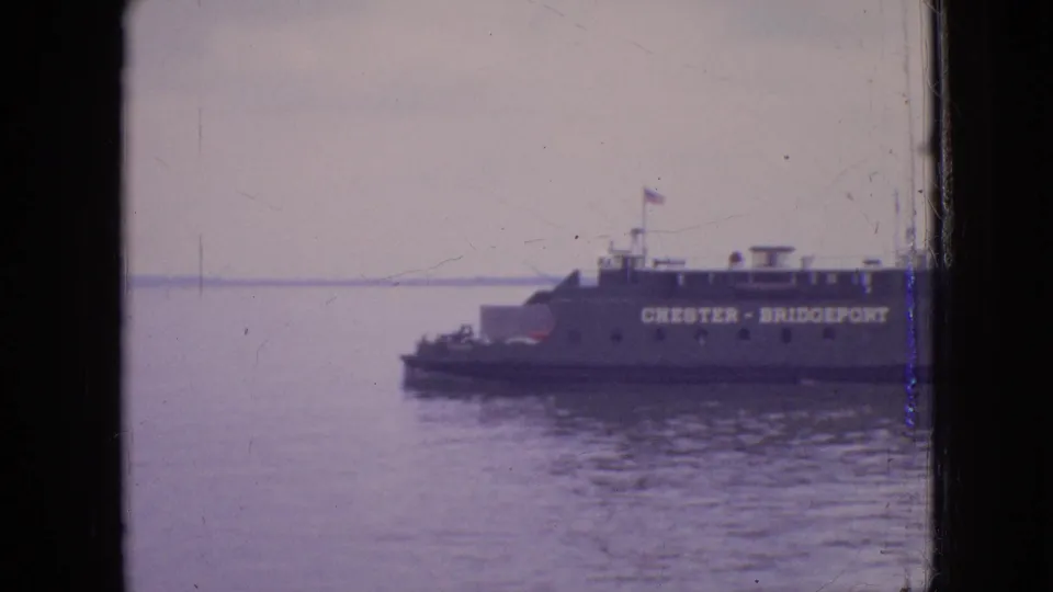 1964 Philadelphia-area 8mm frame showing a CHESTER - BRIDGEPORT ferry on calm water, partly divided by a dark window frame with a hazy shoreline beyond.