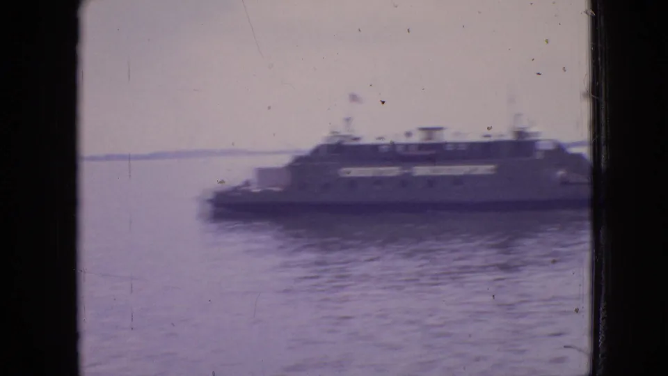Soft 8mm frame of the ferry moving across blue-gray water near Philadelphia, with motion blur, a low shoreline, and dark window edges framing the view.