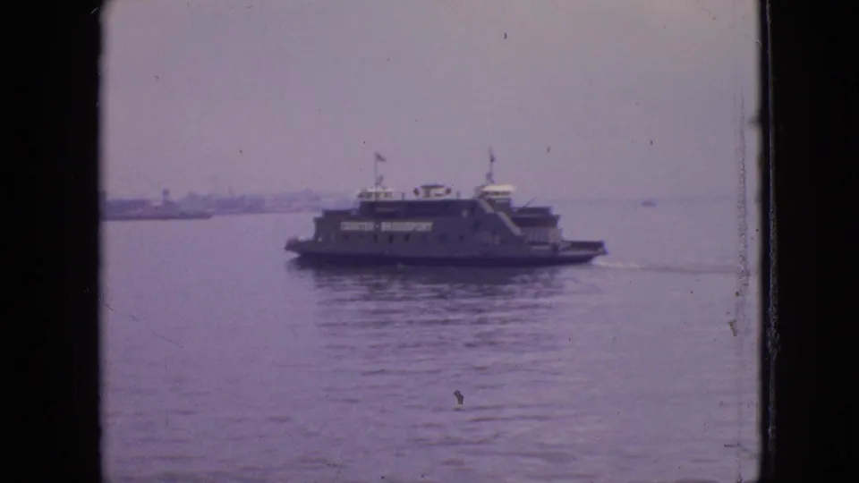 1964 home-movie frame showing the CHESTER - BRIDGEPORT ferry farther across the water, with a small flag, pale sky, and distant shoreline in view.
