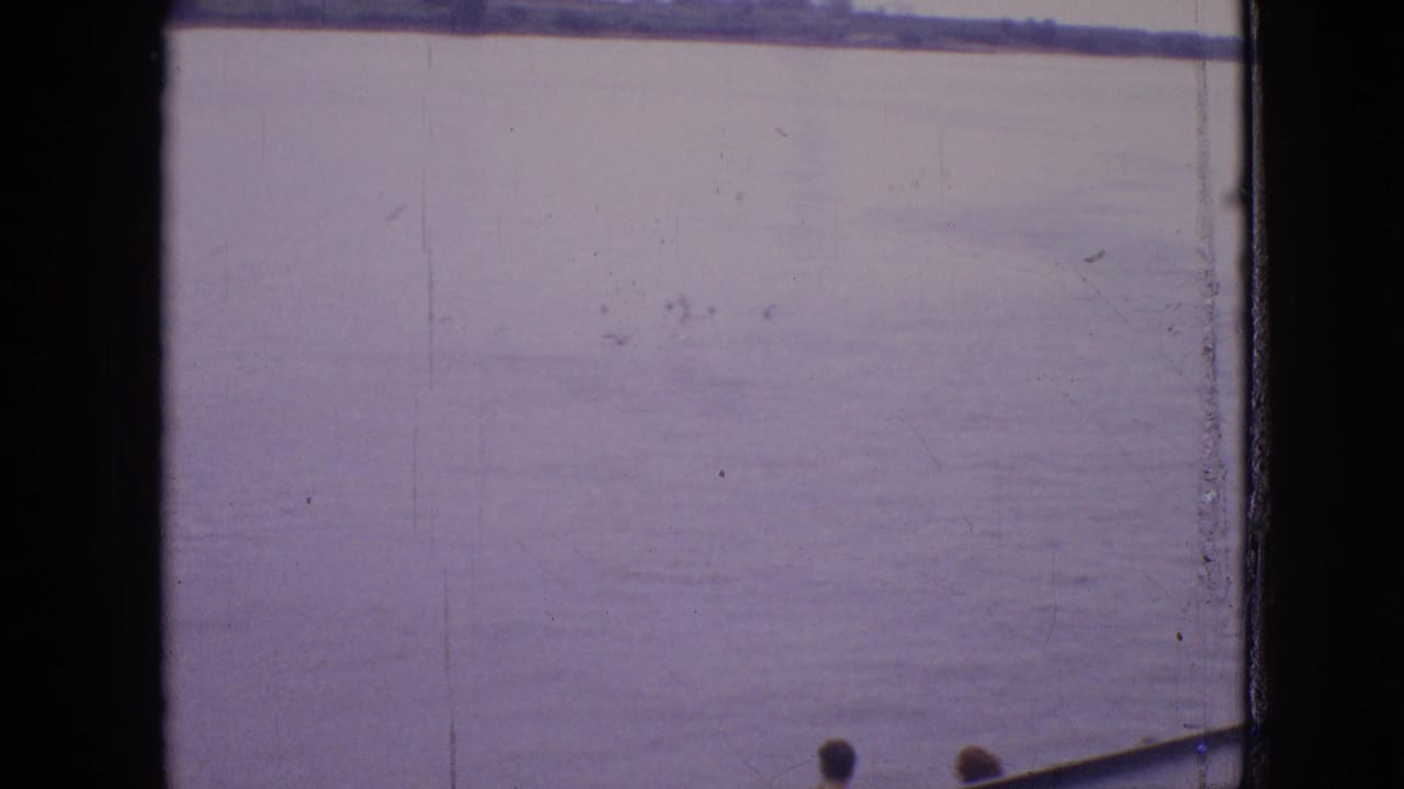 1964 8mm frame showing a calm Philadelphia-area waterway with a distant low shoreline and the edge of a boat entering from the lower right.