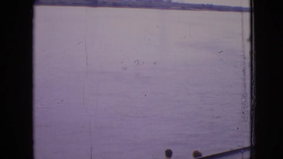 1964 8mm frame showing a calm Philadelphia-area waterway with a distant low shoreline and the edge of a boat entering from the lower right.