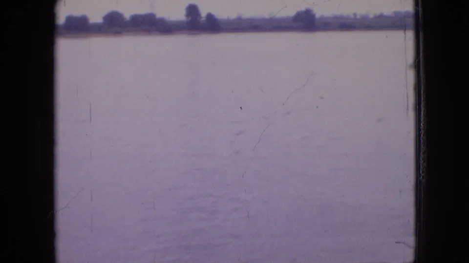 Passenger-view 8mm frame looking across calm water toward a tree-lined shore in the Philadelphia area, with archival scratches and soft color fading visible.