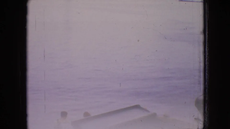 Final 1964 8mm frame from a boat approaching shore, with two partial passenger heads and a boat edge visible against the calm Philadelphia-area water.