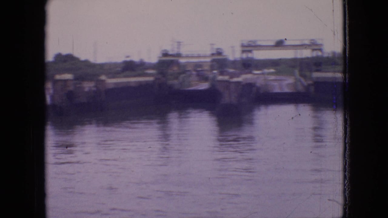 1964 Philadelphia ferry-view frame looking across calm water toward distant industrial pier structures and an overhead gantry-like form.