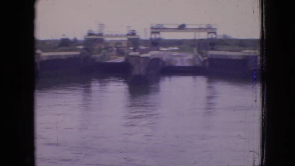 1964 Philadelphia home-movie frame from a ferry crossing, with distant pier structures and a gantry-like form reflected on the water.