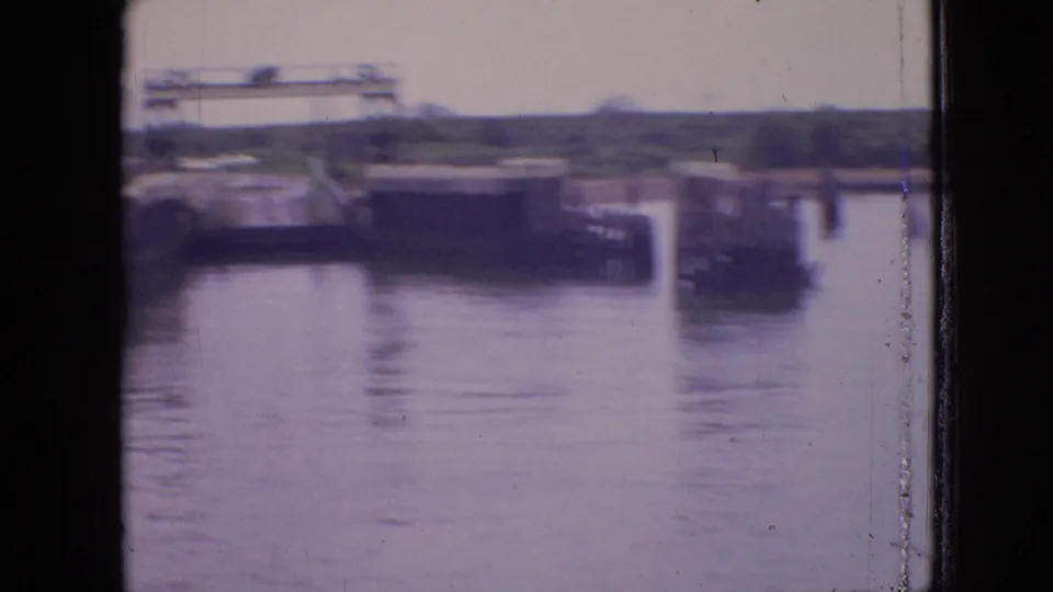 1964 Philadelphia ferry-view frame showing calm water, a distant pier, an overhead gantry-like structure, and a small dark vessel or dock shape near the right edge.