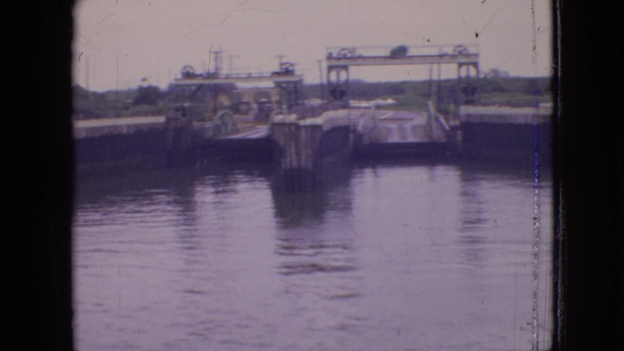 1964 Philadelphia 8mm frame showing a canal lock or water-control passage with calm water, concrete side walls, pilings, and a raised bridge or gate structure in the distance.