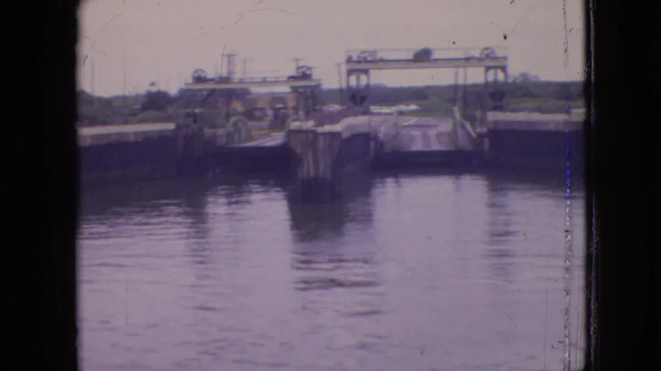 1964 Philadelphia 8mm frame showing a canal lock or water-control passage with calm water, concrete side walls, pilings, and a raised bridge or gate structure in the distance.