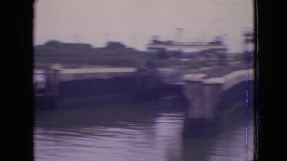 Motion-blurred 1964 home-movie frame showing the side of a canal lock or bridge structure beside water, with concrete edges and low vegetation beyond.