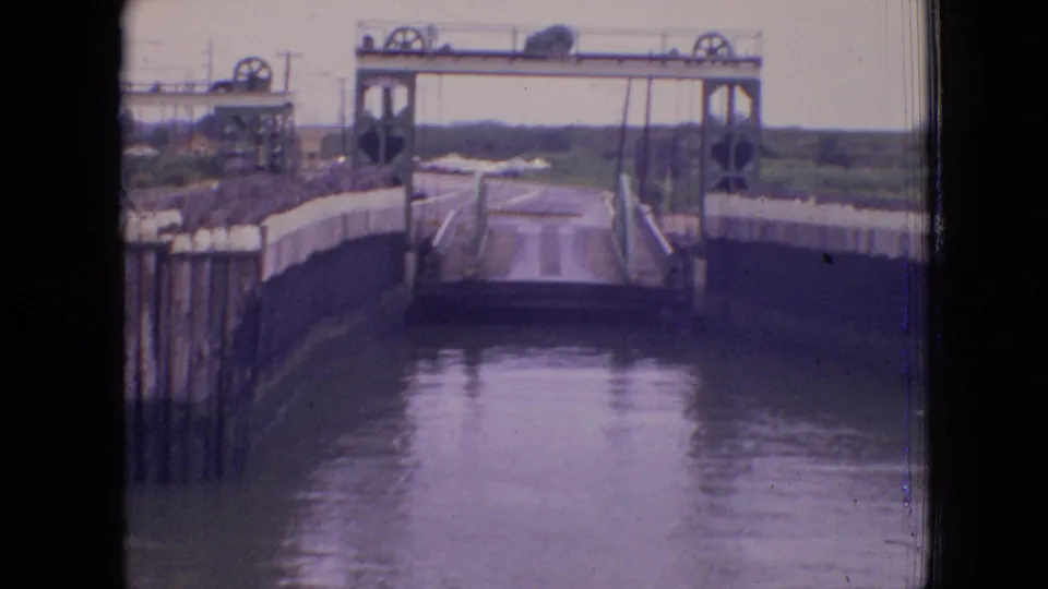 Clearer 1964 Philadelphia 8mm frame of a canal lock or drawbridge area, showing concrete side walls, vertical pilings, railings, mechanical wheels, and water leading through the structure.