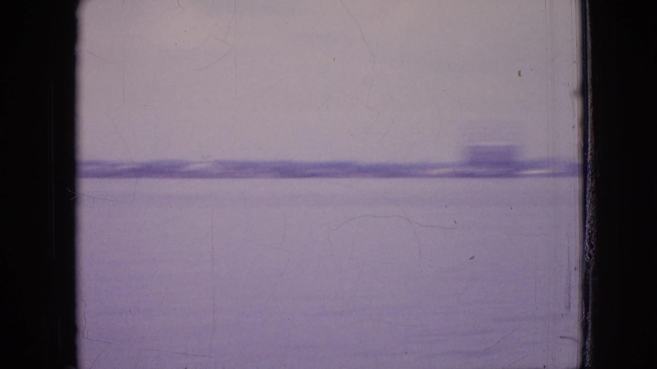 Purple-tinted 1964 8mm view of a distant shoreline across water in the Philadelphia-area clip, framed by dark scan edges.