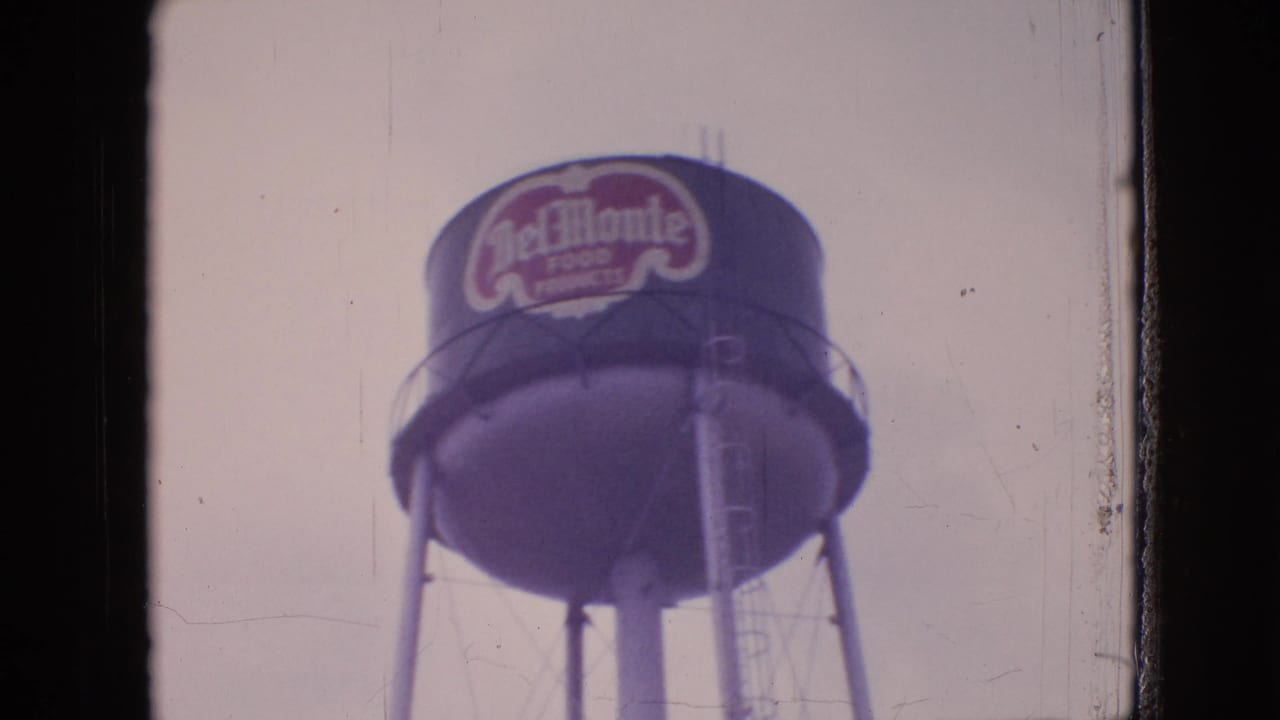 Soft 8mm frame of a Del Monte Food Products water tower rising above an industrial waterfront setting in the 1964 Philadelphia-area footage.