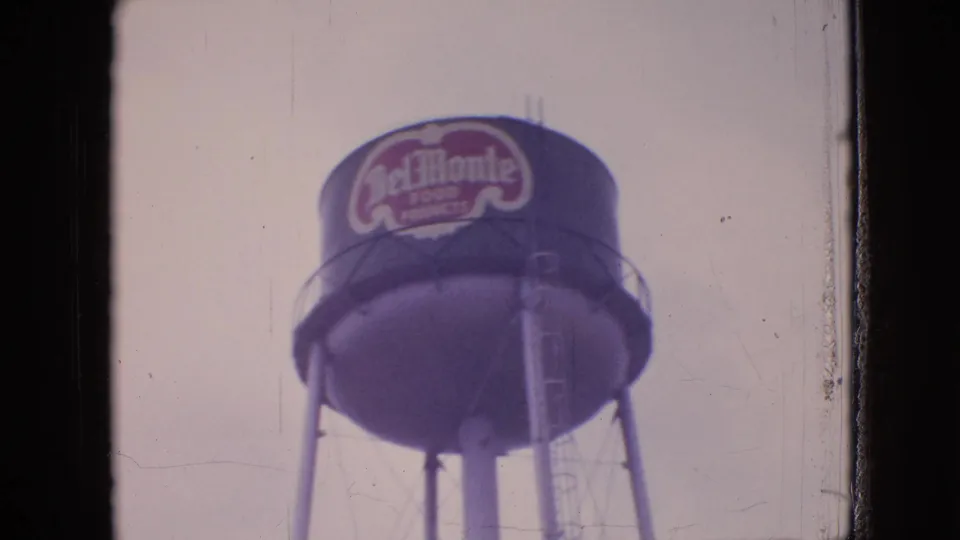 Soft 8mm frame of a Del Monte Food Products water tower rising above an industrial waterfront setting in the 1964 Philadelphia-area footage.