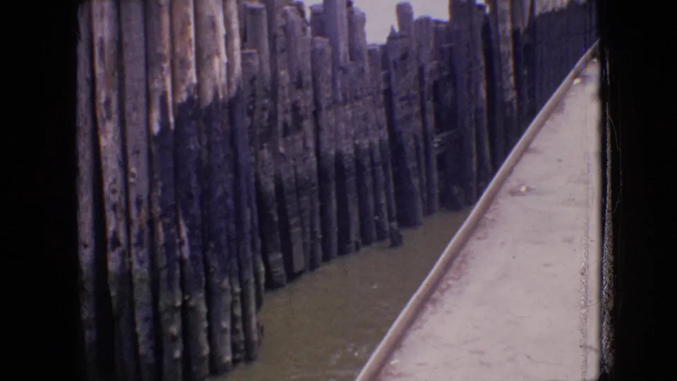 Purple-shifted 8mm view of weathered wooden pier pilings and a narrow concrete walkway along water in the 1964 Philadelphia-area clip.