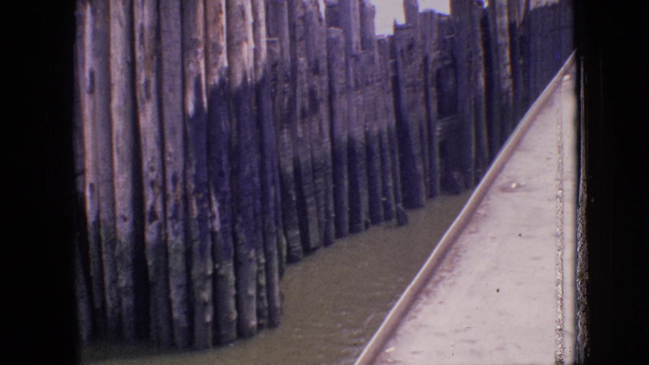 1964 Philadelphia home-movie frame showing tall weathered shoreline pilings beside calm water, filmed from the edge of a moving vessel.