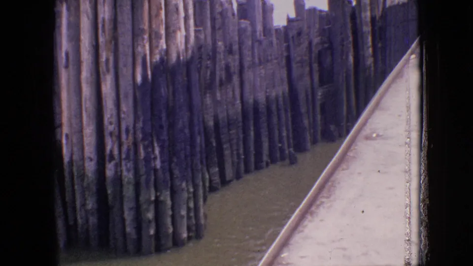 1964 Philadelphia home-movie frame showing tall weathered shoreline pilings beside calm water, filmed from the edge of a moving vessel.
