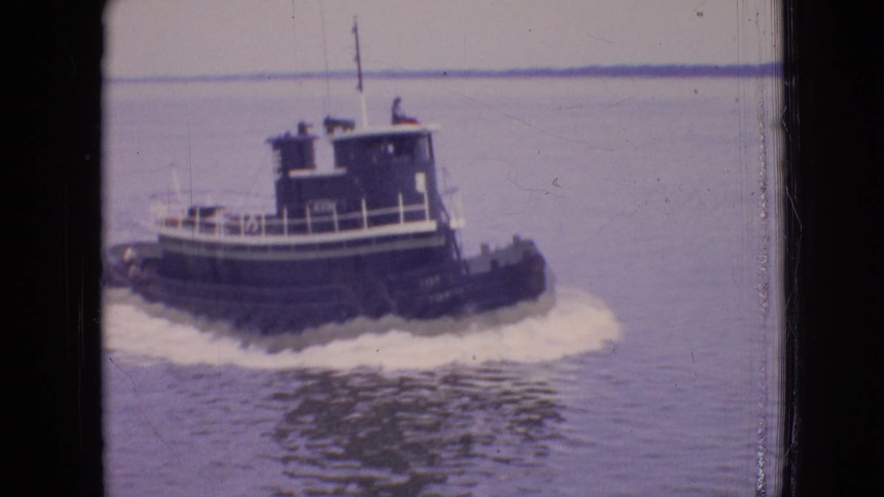 1964 Philadelphia home-movie frame of a dark tugboat crossing calm water with a white wake and a low distant shoreline behind it.
