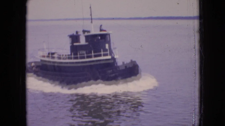 1964 Philadelphia home-movie frame of a dark tugboat crossing calm water with a white wake and a low distant shoreline behind it.