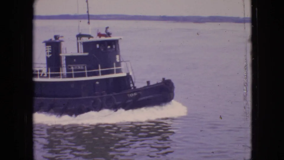 1964 Philadelphia home-movie frame showing a dark tugboat passing close to the camera, with deck railings, cabin, water, and wake visible.