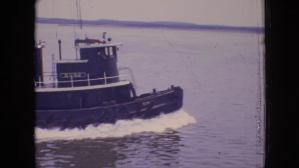 1964 Philadelphia home-movie frame showing the tugboat in partial profile on open water, with a small crew figure near the cabin and wake along the hull.