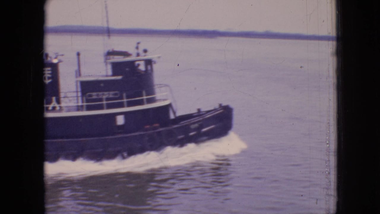 A dark blue or black tugboat with white trim moves across calm Philadelphia harbor water in supplied 1964 8mm home-movie footage, with shoreline faintly visible behind it.