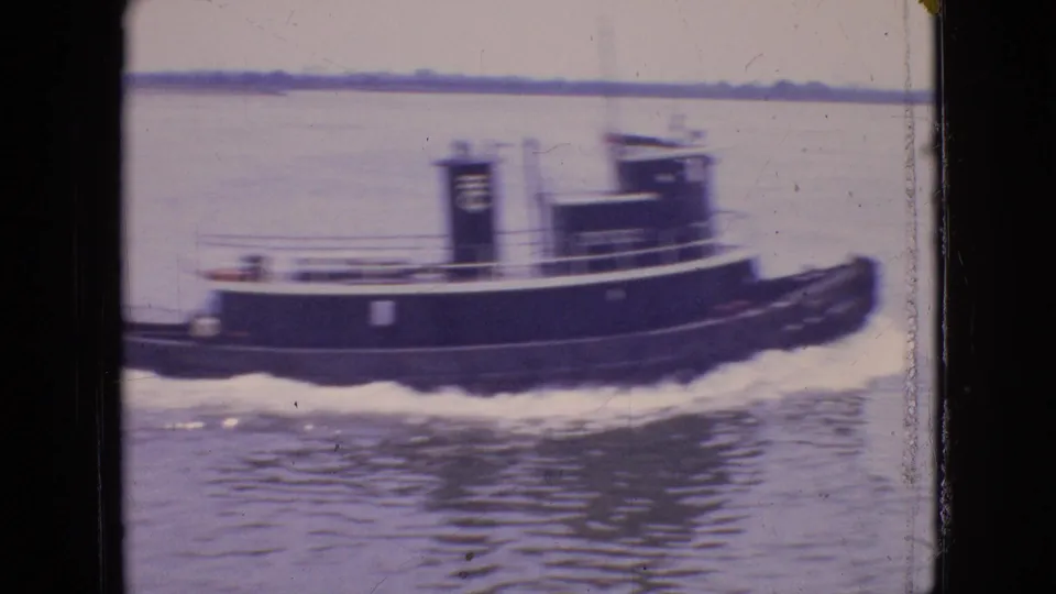 The tugboat continues across the frame on calm water, showing its cabin, railings, dark hull, pale trim, and wake in 1960s home-movie footage identified as Philadelphia.