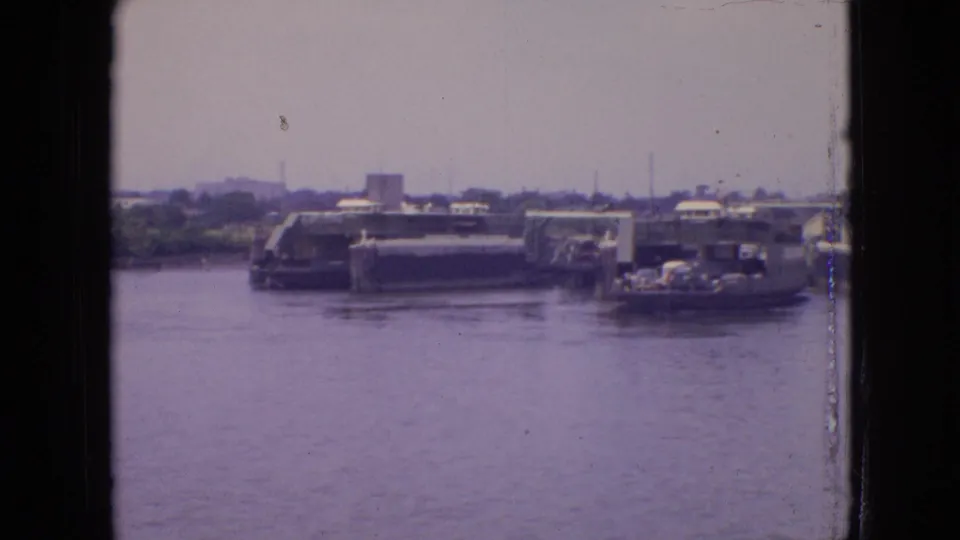 A ferry or docked vessel in the Philadelphia harbor area appears with vehicles visible on or near it, set against low industrial waterfront buildings in the supplied 1964 footage.