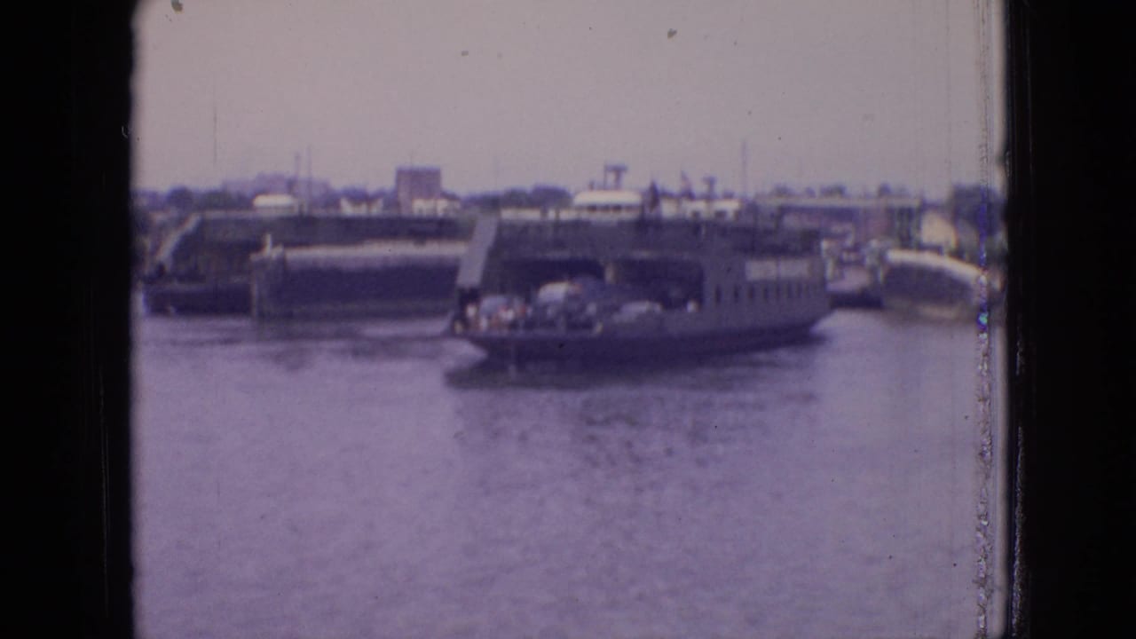 1964 Philadelphia-area 8mm frame showing the Chester-Christopher ferry at a distance with cars aboard, harbor water, and a hazy waterfront behind it.