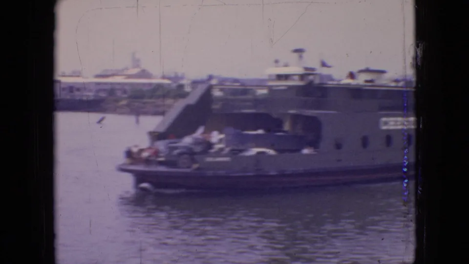 1964 Philadelphia-area 8mm frame showing the Chester-Christopher ferry closer to the camera, with automobiles on deck, passengers near the rail, and a hazy waterfront beyond.