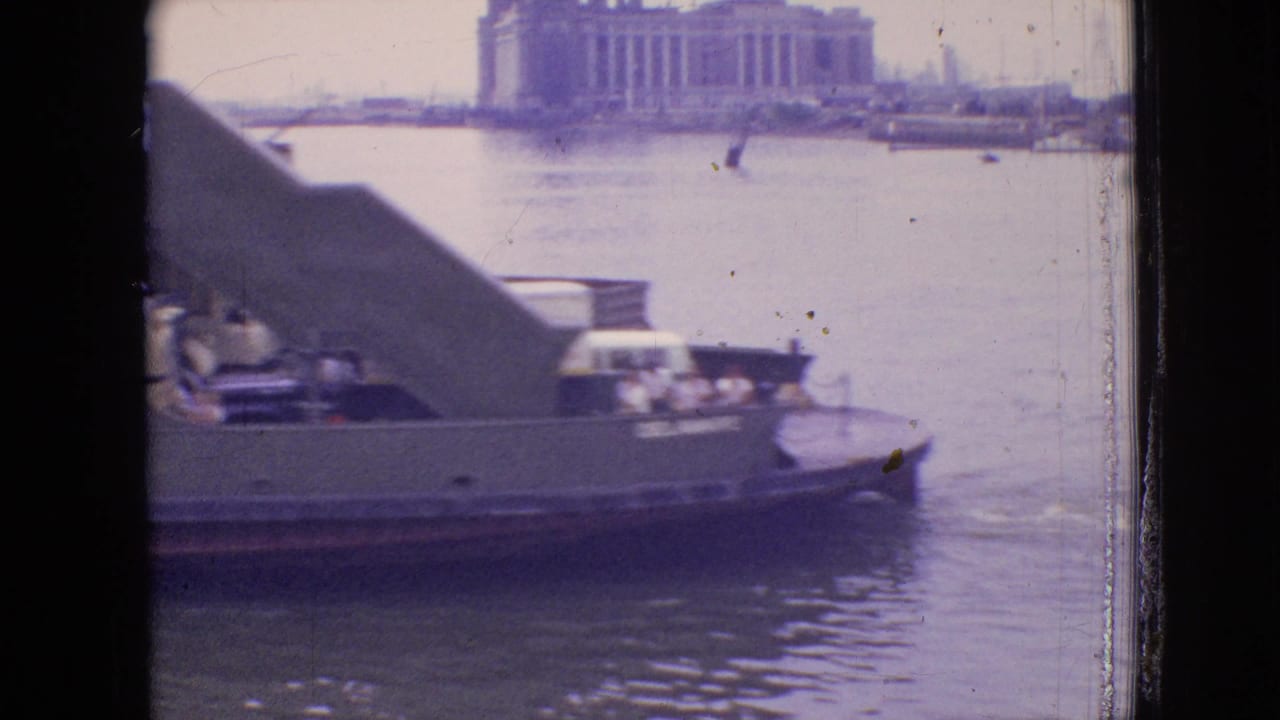 Ferry moving on the water with an open ramp and large building in the background.
