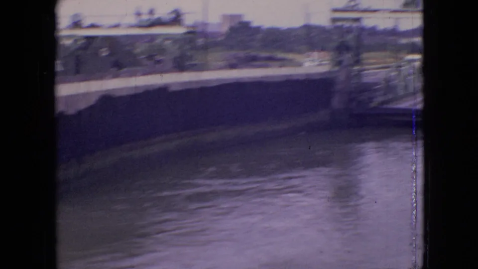 1964 Philadelphia-area ferry footage frame showing the vessel passing close to camera, with blurred passengers, water, and shoreline structures behind it.