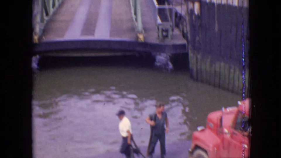 1964 Philadelphia home-movie frame showing two adult men near the water, a red vehicle at the edge of the frame, and a raised green bridge over the canal.