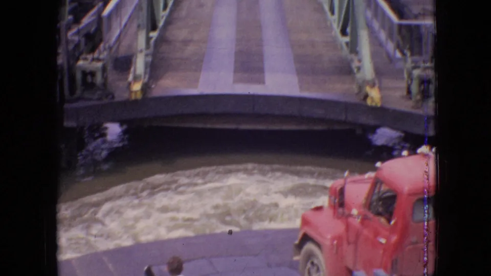1964 Philadelphia frame showing a red vehicle by churned water beneath a raised metal bridge, with a person partly visible at the lower edge.