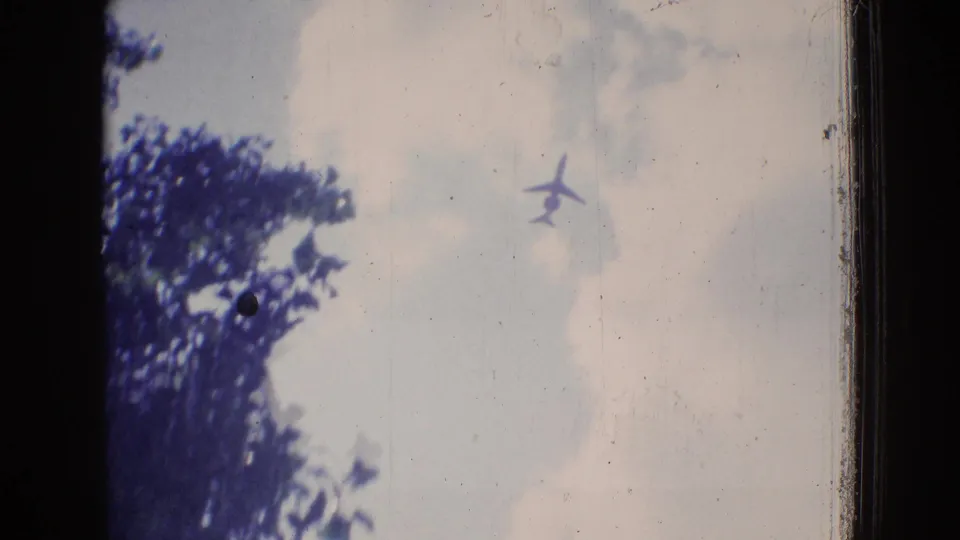 A 1965 New Jersey home-movie frame shows a small airplane silhouette in a pale cloudy sky, with dark tree foliage and visible film wear along the edges.