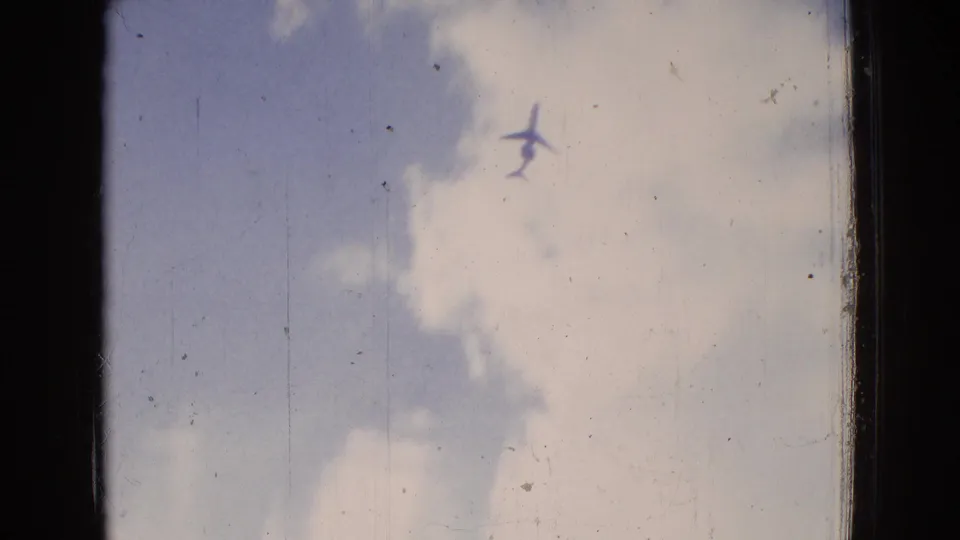 The airplane remains visible against a pale cloudy New Jersey sky in this 1965 8mm frame, with dark side borders, light scratches, and faded blue color.