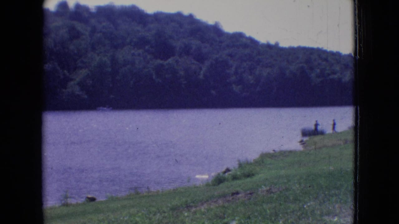 1965 New Jersey 8mm frame showing a calm lake, tree-covered opposite hillside, grassy bank, and small distant people near the shoreline.