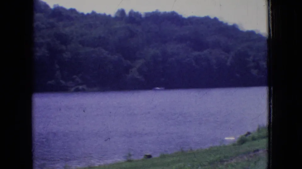 1965 New Jersey 8mm frame looking across a lake toward a dense wooded hillside, with grassy shoreline visible along the right foreground.