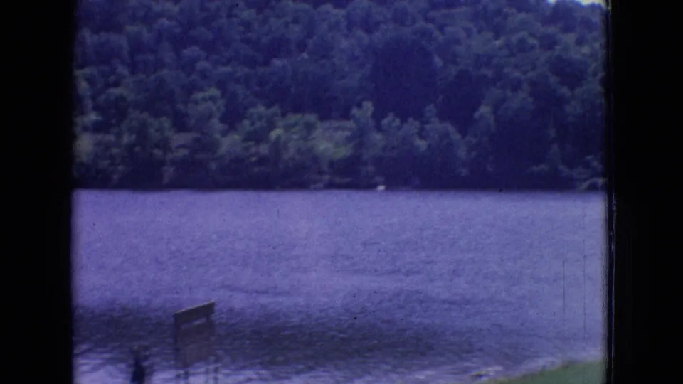 1965 New Jersey 8mm frame showing lake water, a distant wooded shoreline, and a small foreground sign or post near the grassy edge.