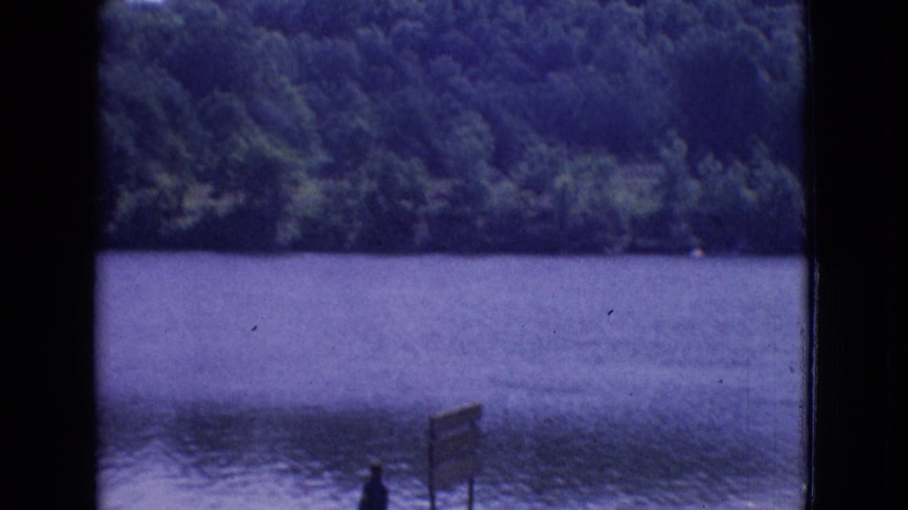 1965 New Jersey 8mm frame showing a calm lake, dense trees across the water, a small standing figure near the shore, and a wooden signpost at the lower edge.