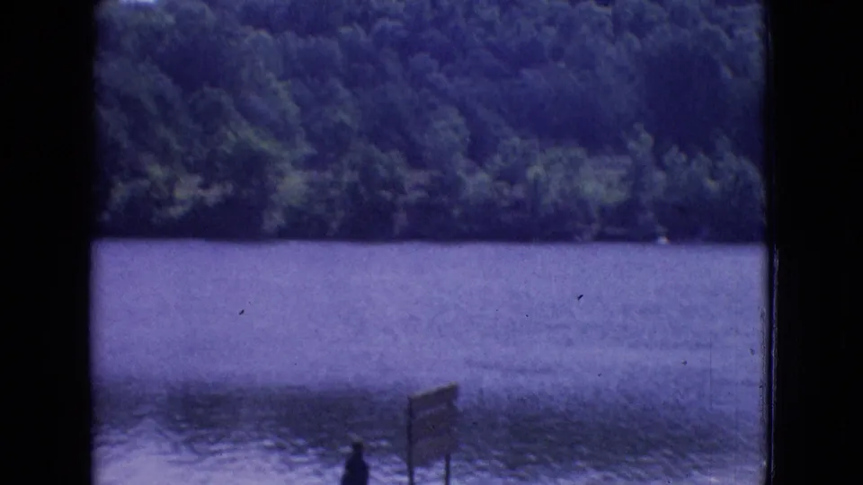 1965 New Jersey 8mm frame showing a calm lake, dense trees across the water, a small standing figure near the shore, and a wooden signpost at the lower edge.