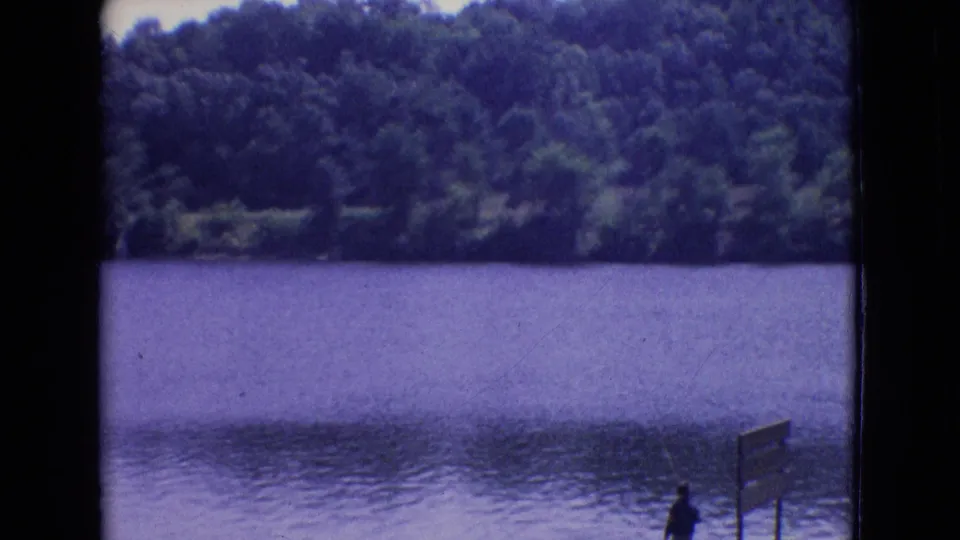 1965 New Jersey 8mm frame of a blue-toned lake and tree-covered shoreline, with a distant person and wooden signpost partly visible in the foreground.