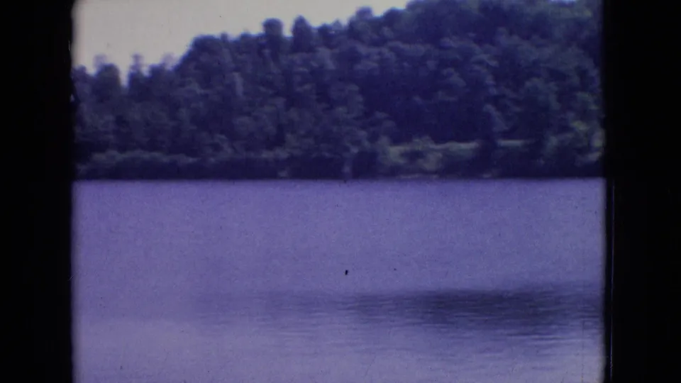 1965 New Jersey 8mm frame showing a tranquil lake, wooded far bank, pale sky, and visible film border with no clearly identifiable foreground person in this frame.