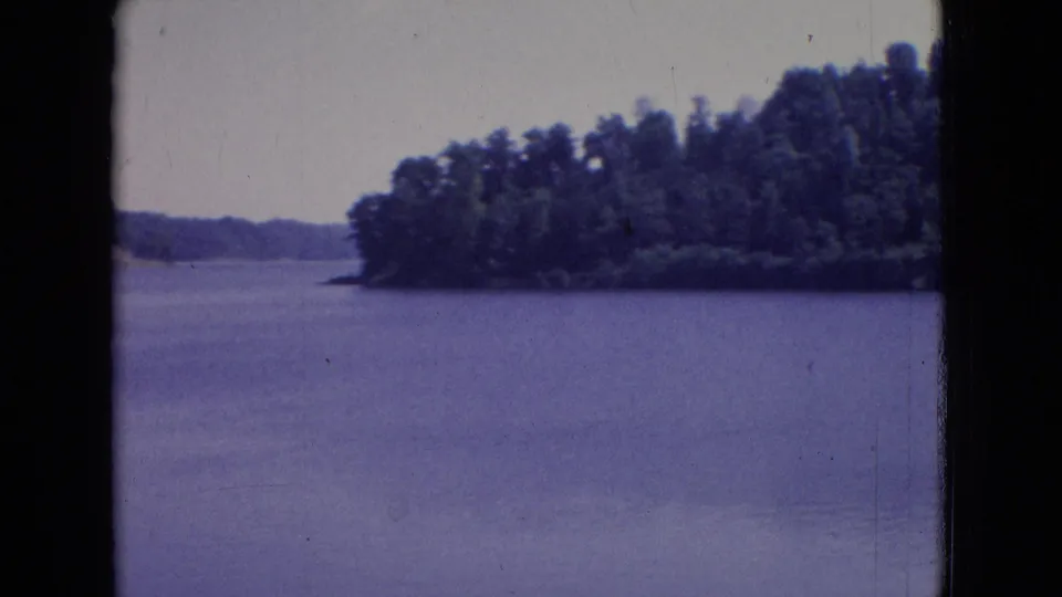 1965 New Jersey 8mm frame looking across a calm lake toward a wooded point of land, with distant shoreline, pale sky, and blue-purple film color cast.