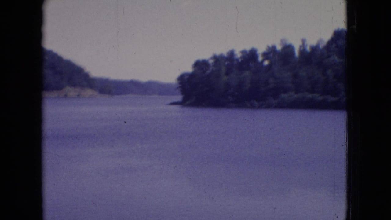 1965 New Jersey 8mm frame showing calm lake water, tree-covered shoreline, distant hills, pale sky, and dark film gate borders.